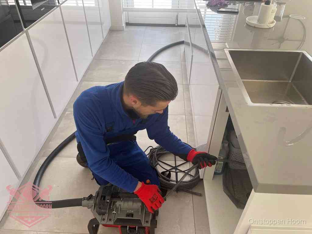 Een monteur ontstopt de keukenafvoer onder de spoelbak met een elektrische veermachine in Hoorn