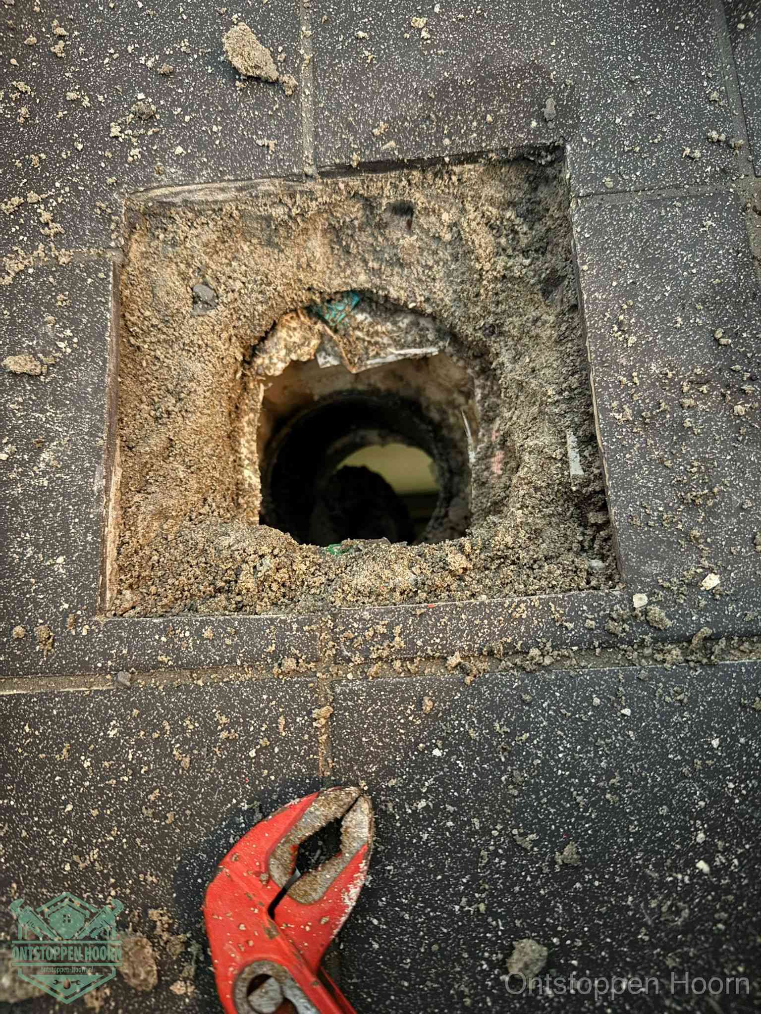 Een geopend rioolputje in een betontegelvloer met losgemaakt rooster en een zichtbare ronde afvoerbuis, het achterhalen waar de afvoerverstopping zit in Hoorn