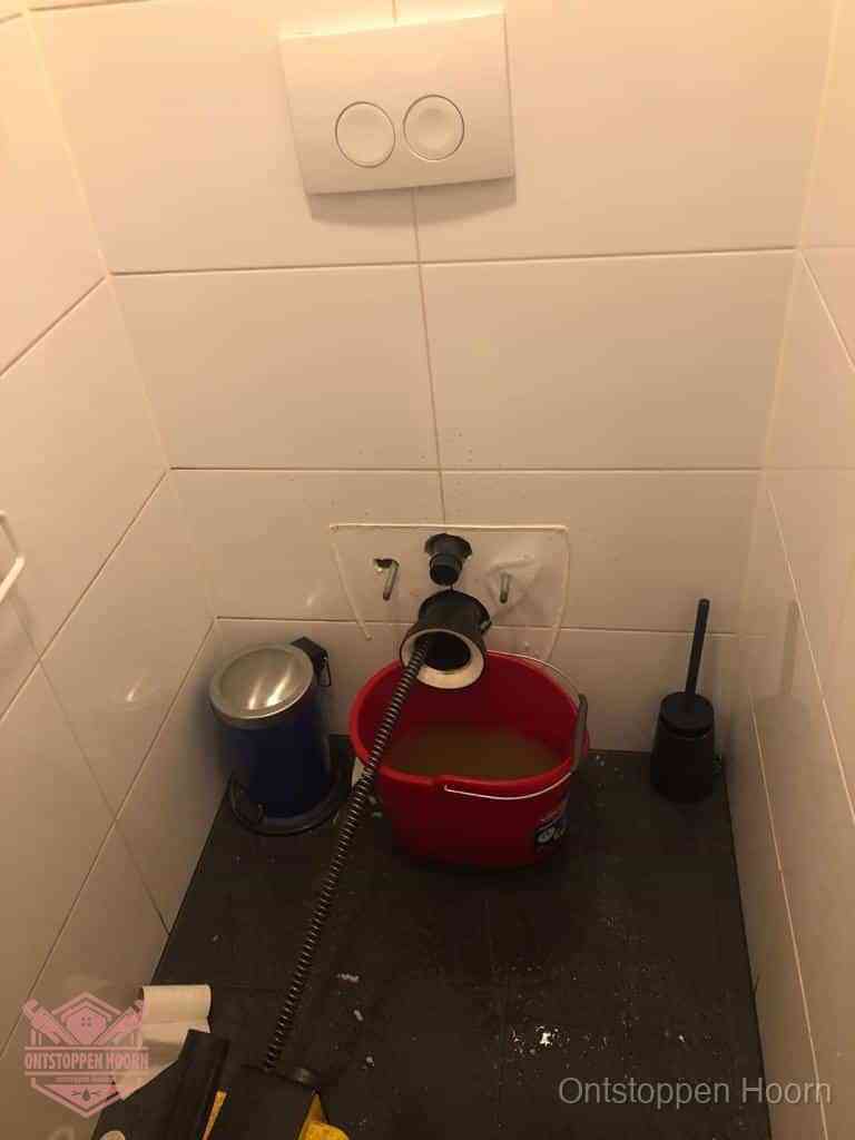 Een ontstopte wand-wc met verwijderde toiletpot waar een ontstoppingsveer in de wanduitgang verdwijnt, het achterhalen waar de verstopping zit in Hoorn