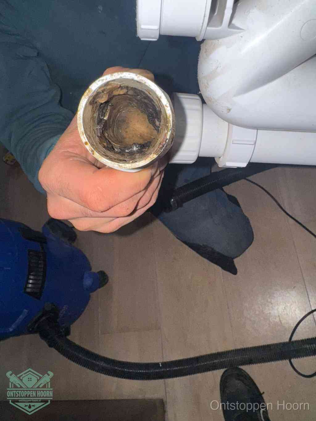 Een hand houdt een losgekoppeld PVC-afvoerbuisstuk vast dat van binnen volledig is dichtgeslibd met vet, vuil en aanslag, de meest voorkomende oorzaak van een verstopte afvoer in Hoorn