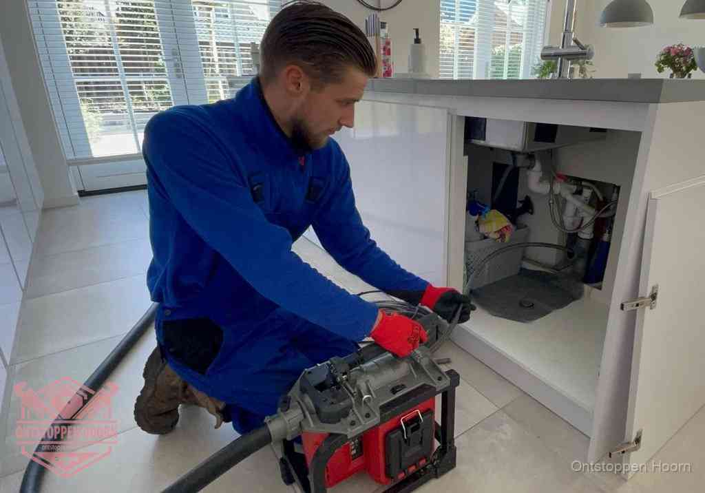 Een monteur voert een professionele elektrische ontstoppingsmachine in de afvoer onder de spoelbak tijdens het gootsteen ontstoppen in Hoorn